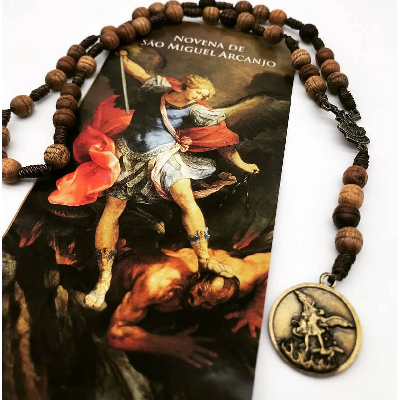 Mãos rezando o terço diante de imagem de São Miguel Arcanjo